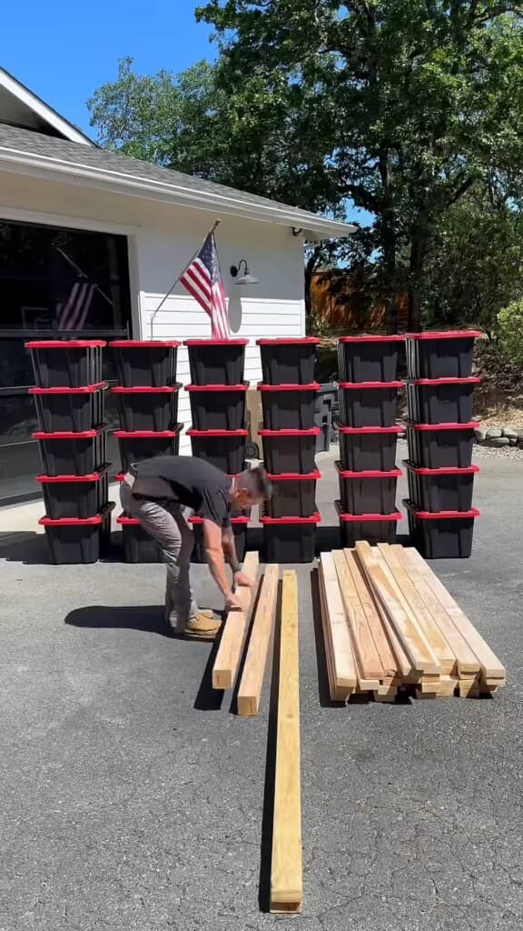 地面に多数の木材を並べて準備する男性。後方には収納ボックスが6列に積まれている。青空の下でDIY作業を行っている。