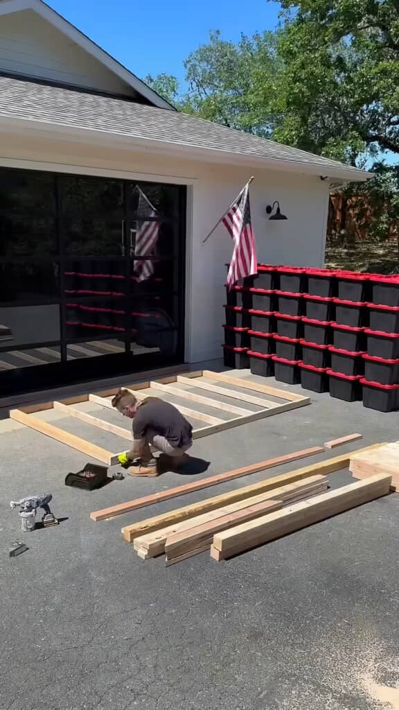 屋外のガレージ前で、男性が木材を床に並べ、収納棚のベースフレームをネジで組み立てている様子。背景にはアメリカ国旗と収納ボックス。