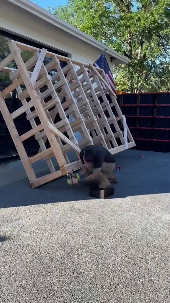 DIY作業中の男性が木製のラックフレームを組み立てている。裏側が見える構造で、ビス留めの作業中。背景には収納ボックスが並んでいる。