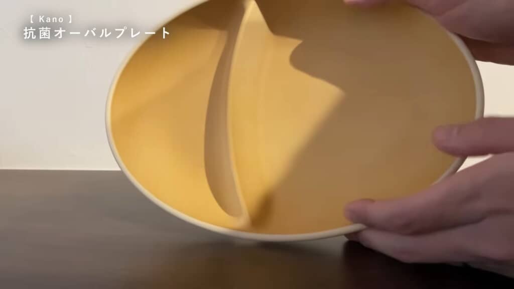 Kanoの抗菌オーバルプレートを手に取っている場面。プレートの色はマットなベージュで、仕切りが中央に入っている。左上に商品名の日本語テキストあり。