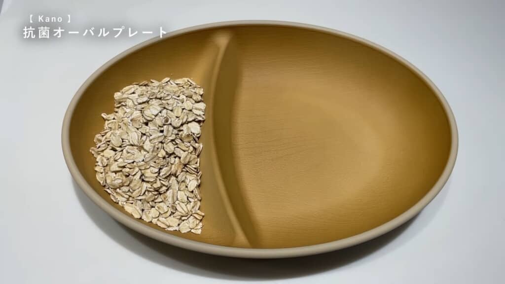 左半分にオートミールを盛り付けた、仕切り付きのベージュ系オーバルプレート。左上に「Kano 抗菌オーバルプレート」という日本語テキスト。