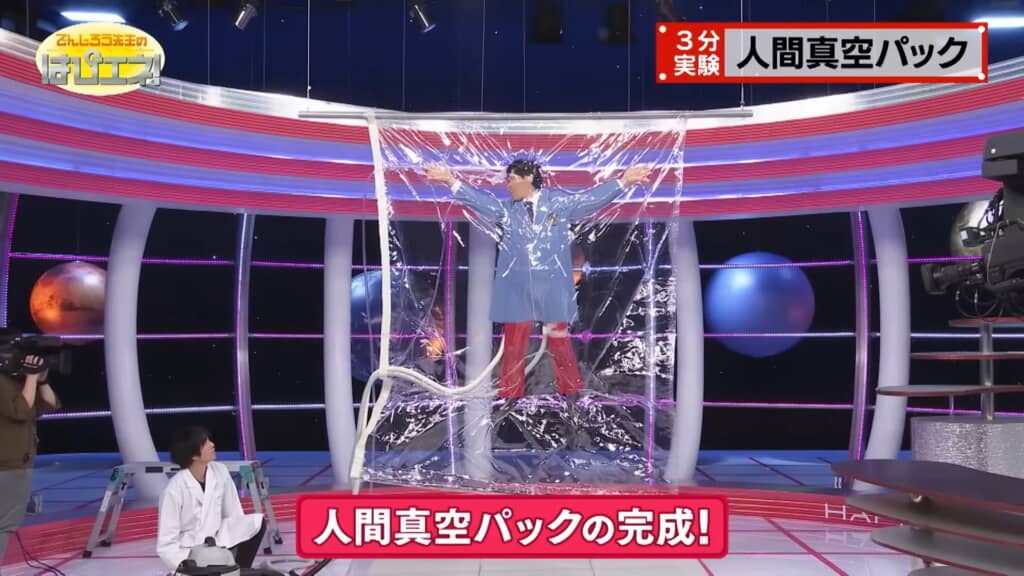 完成した人間真空パックの実験で、男性がX字の姿勢で透明な袋の中に封じられている。袋の外側には掃除機のホースがつながれており、下部には「人間真空パックの完成！」という赤いテロップが表示されている。