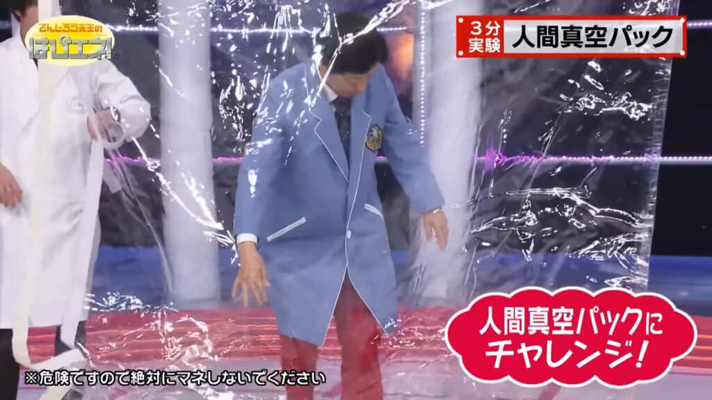 透明なビニールの中に入った男性が、足元の赤いマットの上に立ちながら準備をしている様子。左には白衣姿の人物がホースを持っており、右下には「人間真空パックにチャレンジ！」と赤い吹き出しが表示されている。