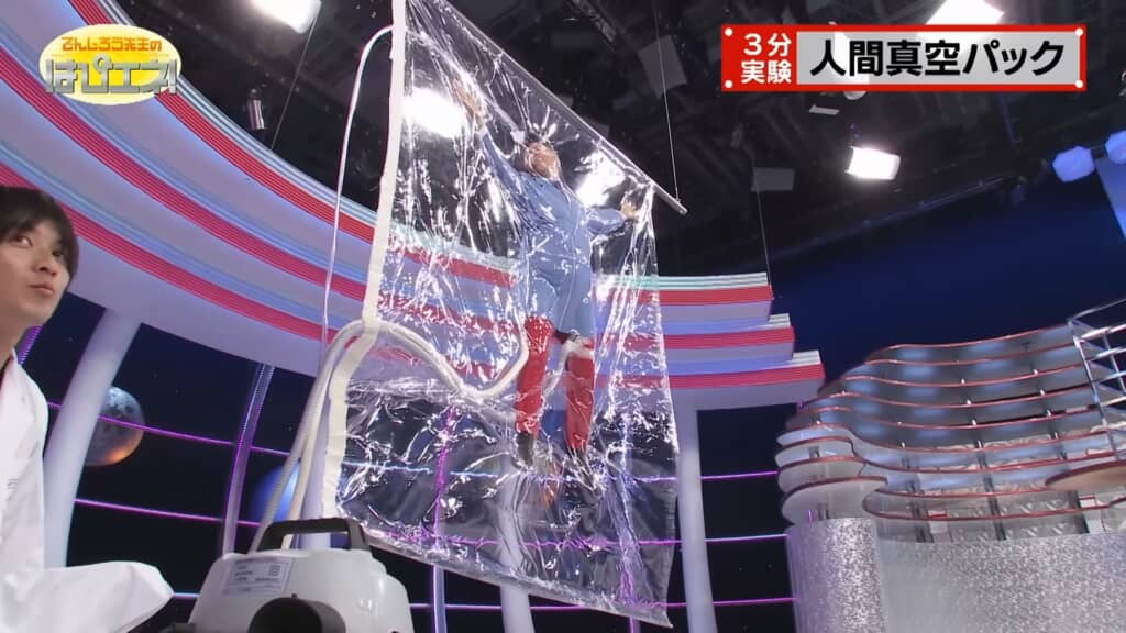 真空パックの実験で、透明なビニール袋の中に立ったまま封じ込められている男性。スタジオのセットにはカラフルなライトが設置され、右上には「3分実験 人間真空パック」と表示されている。
