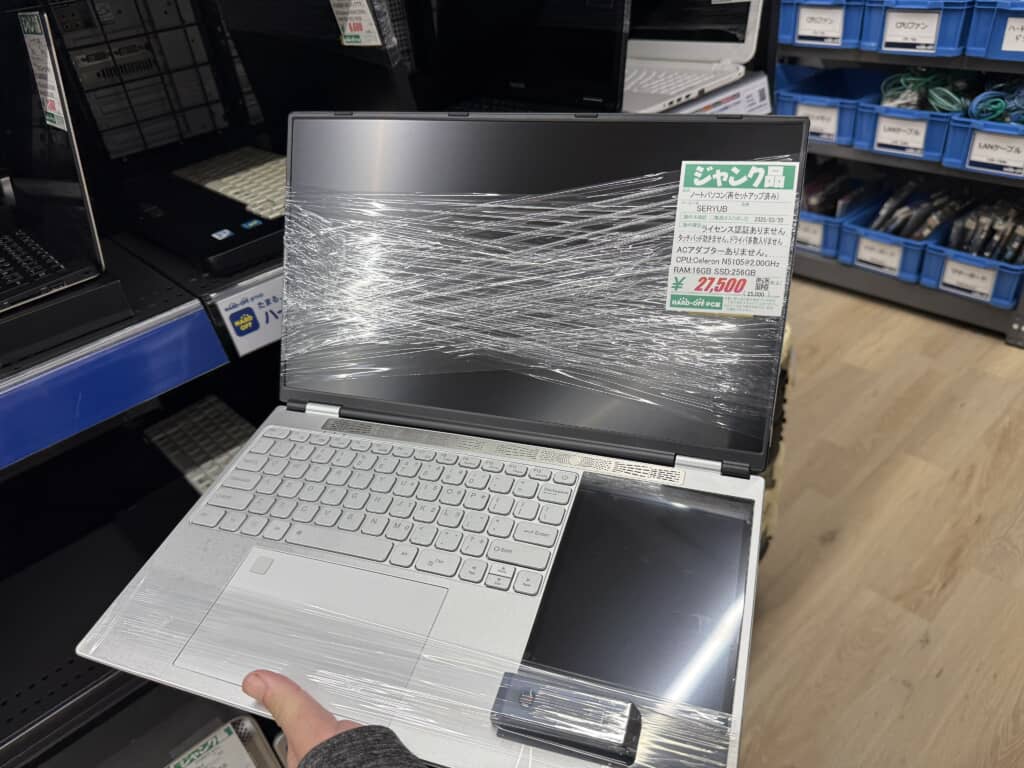 中古パソコン売場で展示されている2画面ノートパソコン。画面と本体は透明ラップで包まれており、右側のセカンドディスプレイが特徴的。ラベルには『ジャンク品』とあり、スペックはCeleron N5105、メモリ16GB、SSD256GB。価格は27,500円（税込）。背景にはLANケーブルなどのパーツが収納された青いボックスが並んでいる。