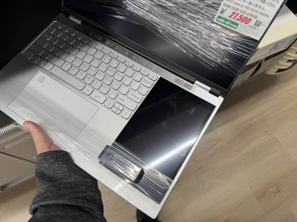 店内で手に持たれている中古ノートパソコン。キーボードとタッチパッド部分は銀色で、右側に黒いサブディスプレイが搭載されている。画面全体がラップで保護されており、値札には『ジャンク品』と明記されている。CPUはIntel Celeron N5105、RAM16GB、SSD256GB、価格は27,500円（税込）。