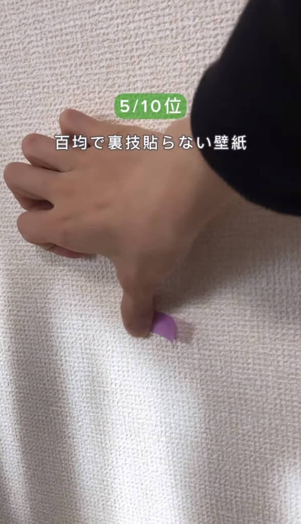 人物の手が壁に貼った壁紙の端を指で押さえているアップショット。紫色の剥がしやすいテープが見える。テキストは『5/10位 百均で裏技貼らない壁紙』。