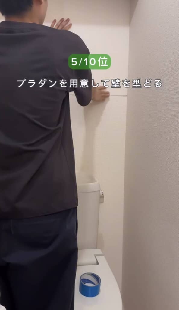 人物がトイレの後方の壁にプラダンをあてて型取りをしている場面。青い養生テープが床に置かれており、DIYの作業途中。テキストは『5/10位 プラダンを用意して壁を型どる』。