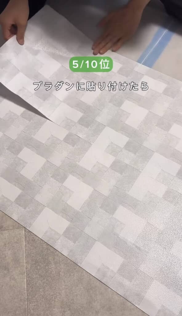 市松模様の壁紙シートを床に広げ、左側の人物の手が壁紙の端を持ち上げて貼り付け作業を行っている様子。中央には『5/10位 プラダンに貼り付けたら』のテキスト。