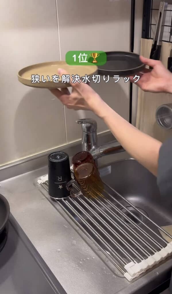 キッチンシンクの上に設置された水切りラックの上に、黒と茶色のマグカップが伏せて置かれている。人の手が2枚の皿をラックに載せようとしている様子。上部に『1位 狭いを解決水切りラック』のテキスト。