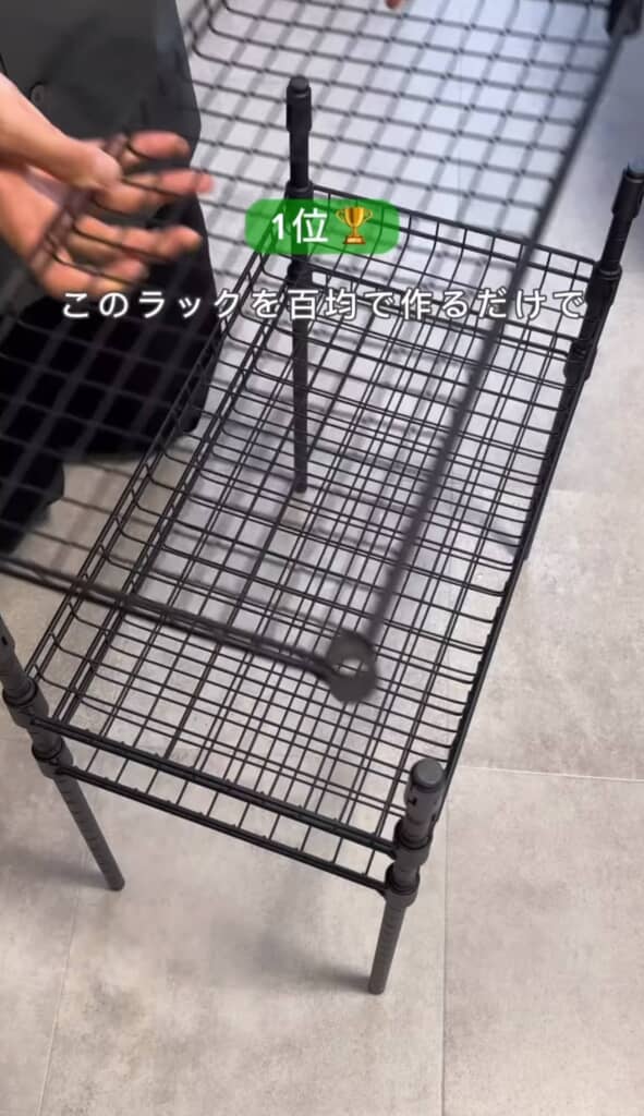 黒いワイヤーラックを手で組み立てている様子。床に設置された下段に追加の網棚を取り付けようとしている。上部には『1位 このラックを百均で作るだけで』というテキスト。