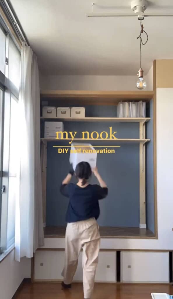 女性が白い収納ボックスを持ち上げて棚に設置している作業中の様子。天井からは1灯の照明が吊り下がっている。