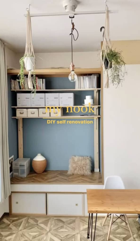 DIYで設置された棚に収納ボックスや雑誌、観葉植物が並ぶ完成後の状態。白い椅子と木製テーブルが前に配置されている。