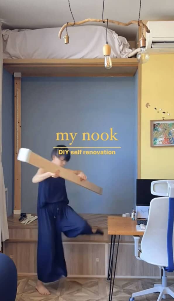 流木の照明が印象的なロフトベッド下でDIYを進める女性。左にはカーテン、右側には木製のデスクとオフィスチェア。