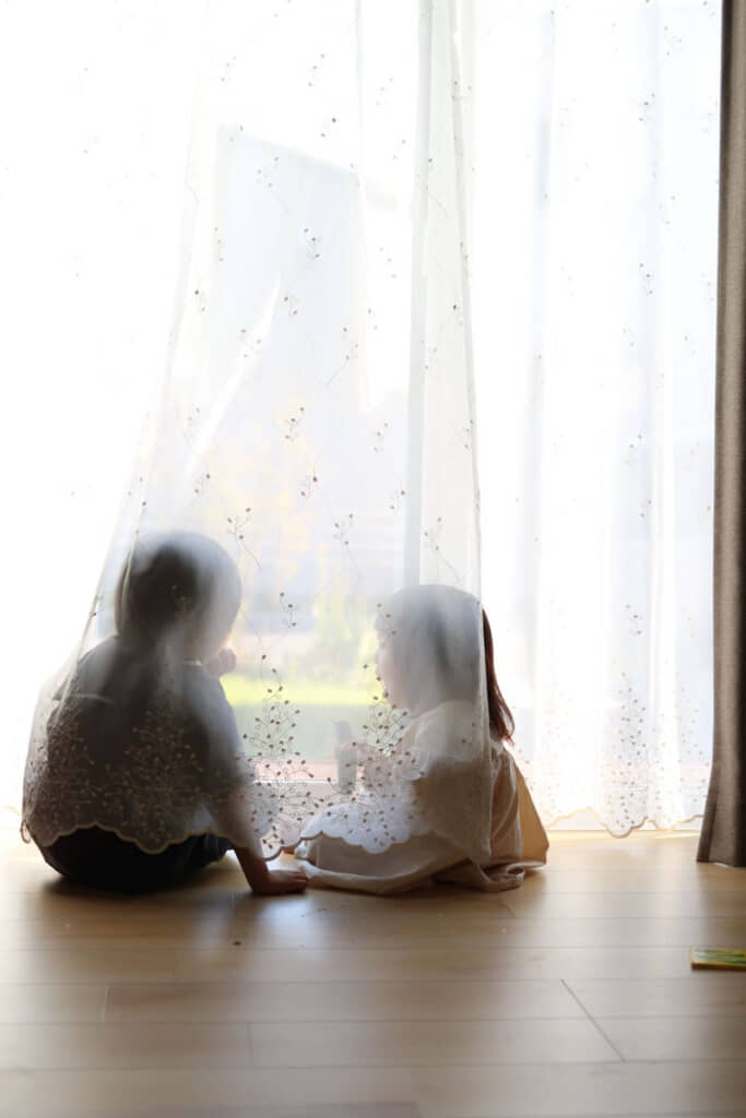 カーテンの向こうで庭を眺めるきょうだい。絵本の挿絵みたい