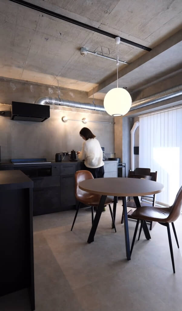 コンクリート打ちっぱなしの天井と無骨な配管が印象的なキッチンで、女性が電気ケトルを操作している様子が写されている。