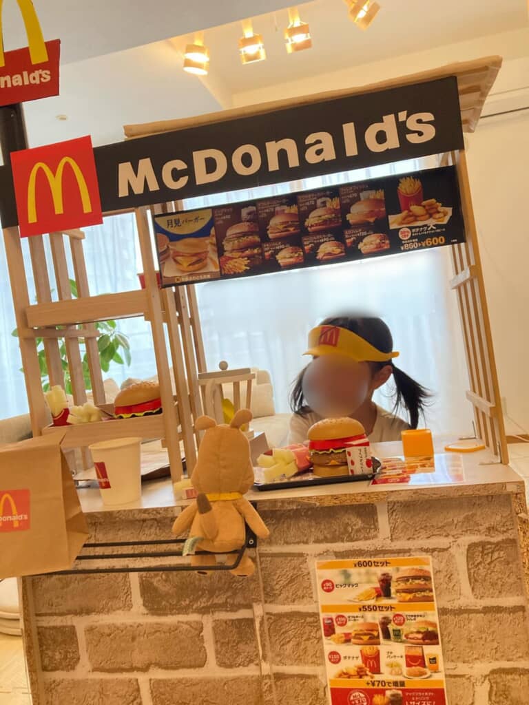 マクドナルド風のごっこ遊びブースで、制服と帽子を身に着けた子どもが接客している様子