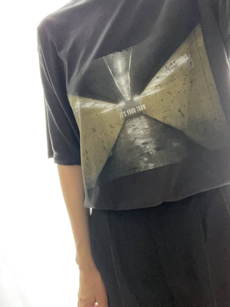 「IT'S YOUR TURN」と書かれたトンネルの写真がプリントされた黒いTシャツを着た人物の上半身。