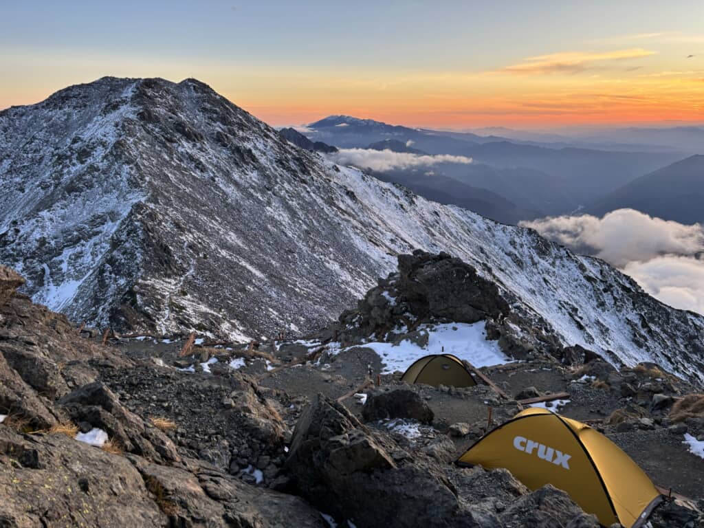 雪の残る山岳地帯に張られたテントと、夕焼けに染まる山並みの絶景