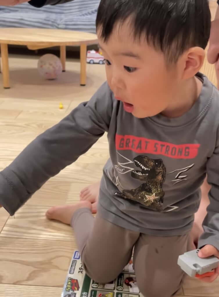 驚いた表情で床に座り、おもちゃを指さす子ども