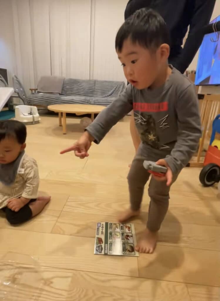 室内で幼児が指を差して立ち、足元の恐竜おもちゃを見て驚いている様子。
