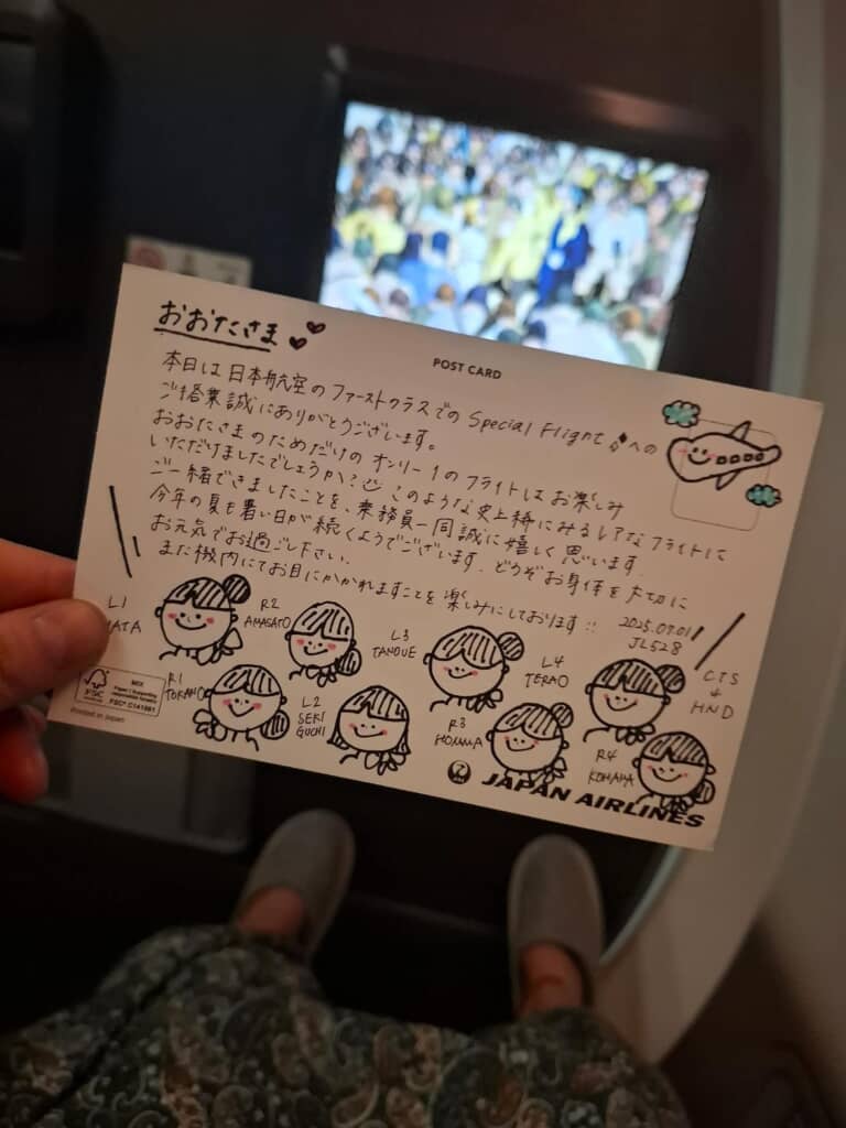 JALの機内で手描きイラスト入りのメッセージカードを手に持ち、前方モニターが映っている様子