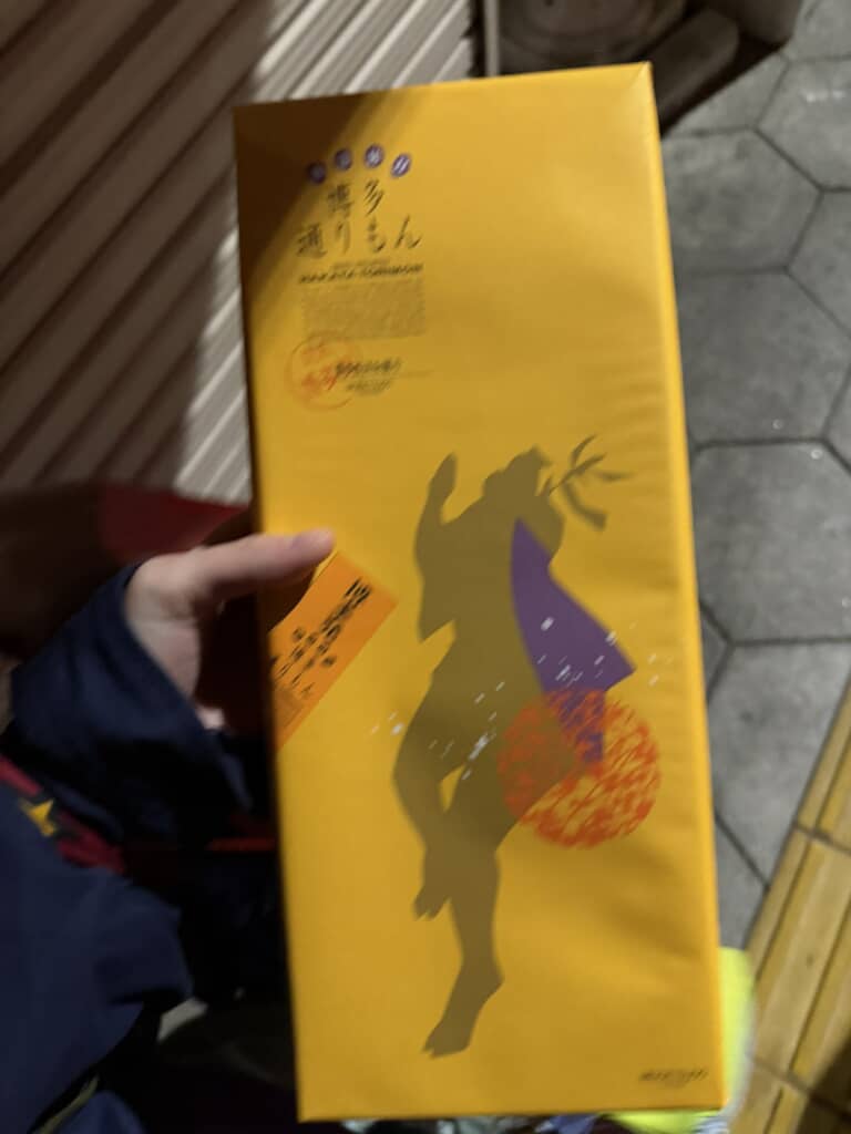 夜の屋外で「博多通りもん」の黄色い包装箱を持つ手元の様子