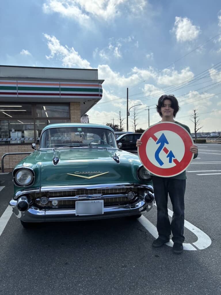 愛車の前で追越し禁止の標識を持つImaiさん
