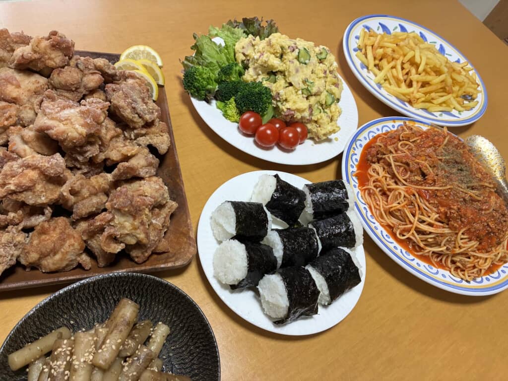 唐揚げ、おにぎり、パスタ、ポテトサラダなどの豪華な家庭料理の盛り合わせ