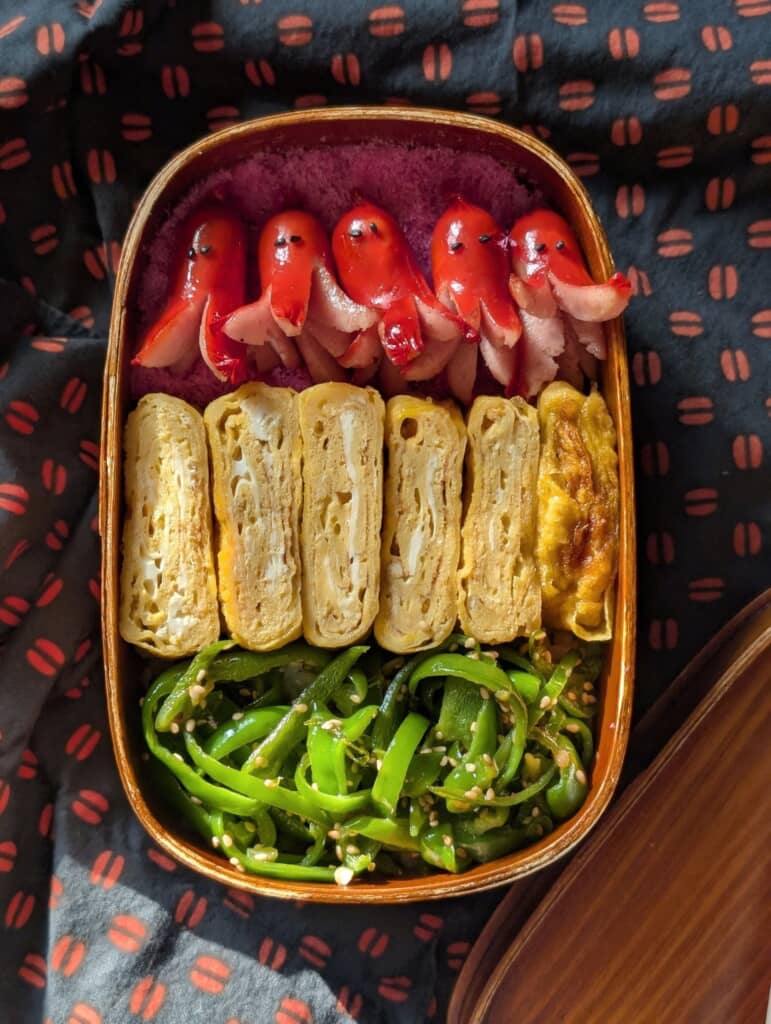 木製弁当箱に並ぶ厚焼き玉子、ピーマンの胡麻和え、タコさんウインナーの弁当の俯瞰
