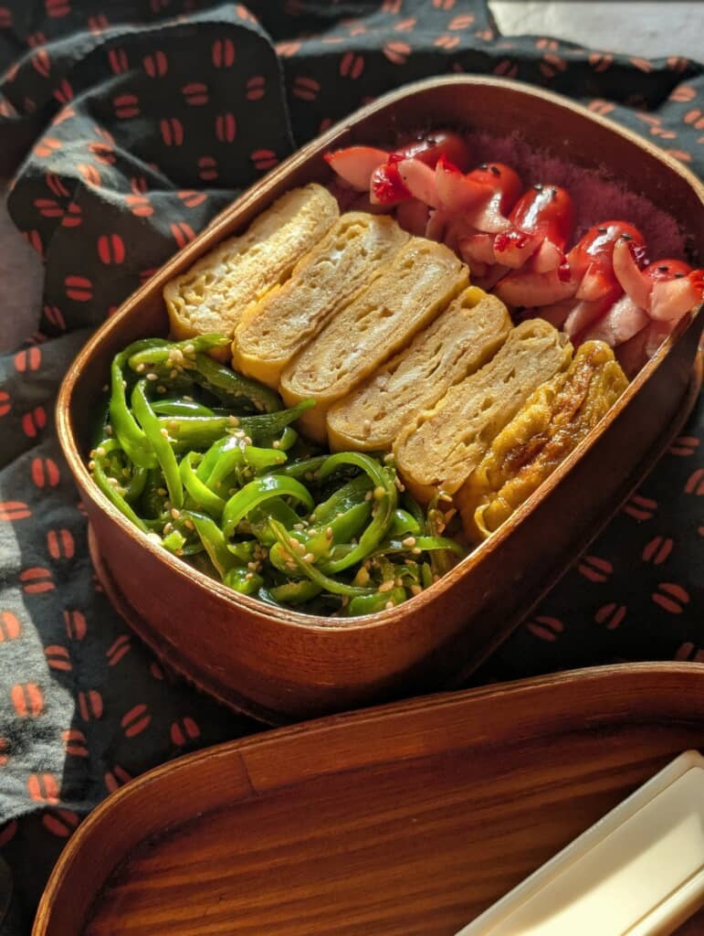 木製弁当箱に詰めた厚焼き玉子、ピーマンの胡麻和え、タコさんウインナーの弁当