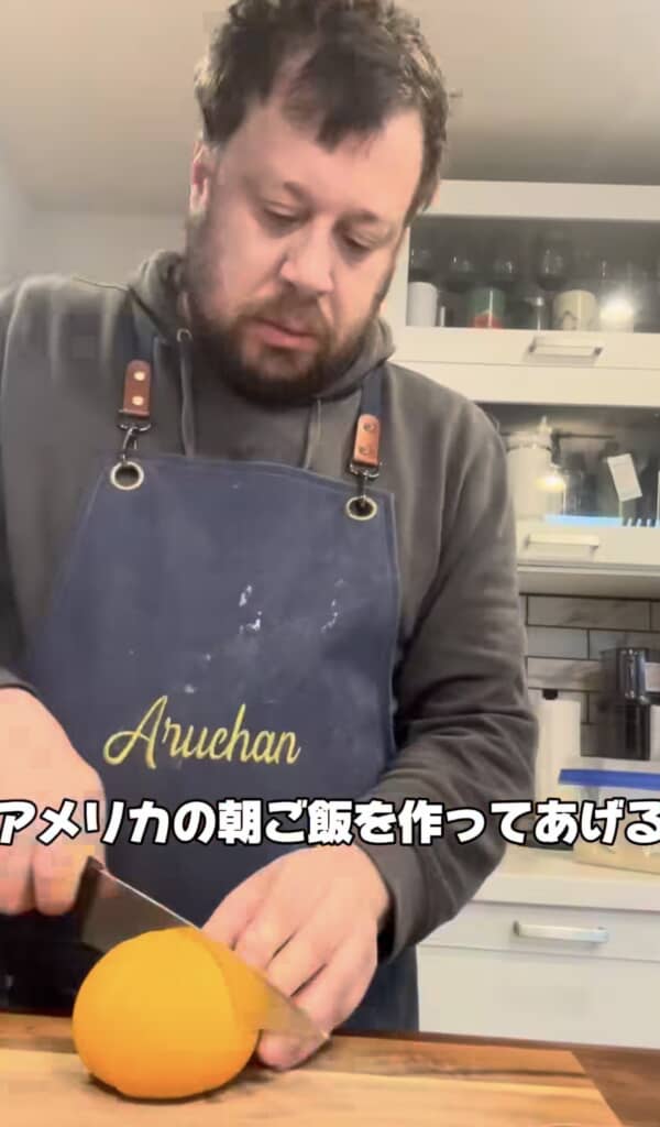"Aruchan"のエプロンをつけた男性がキッチンでオレンジをカットする様子。テロップ「アメリカの朝ご飯を作ってあげる」