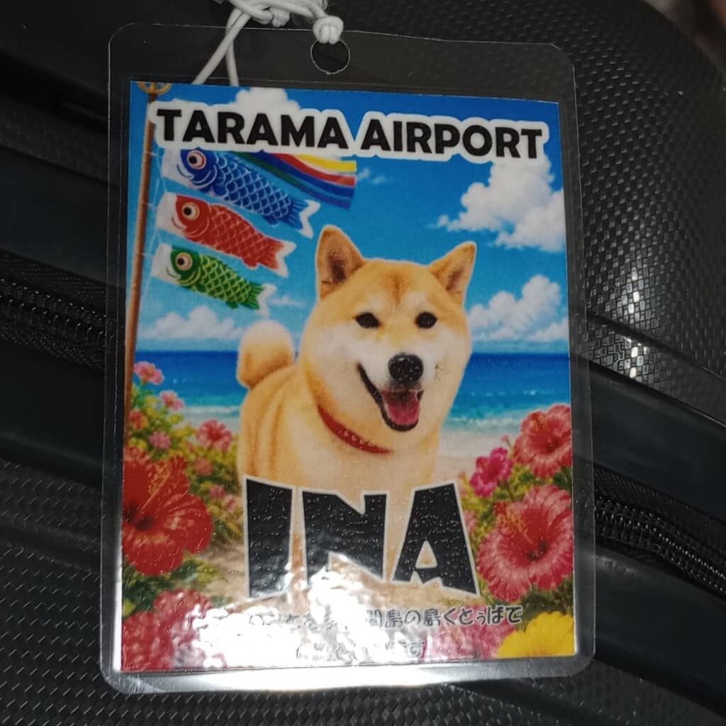 多良間空港のラゲッジタグ「INA」。笑顔の柴犬と青い海・ハイビスカス・こいのぼりのイラスト