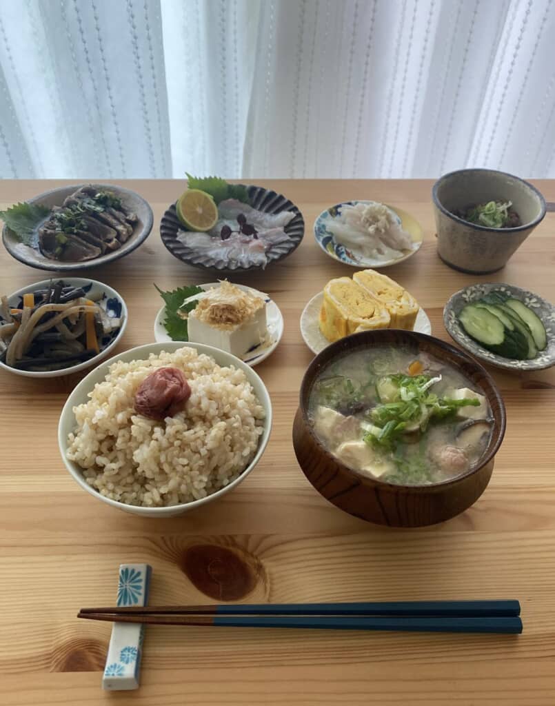 梅干し入り玄米ご飯・具だくさん味噌汁・刺身・だし巻き卵など和食の一汁多菜が並ぶ朝食膳