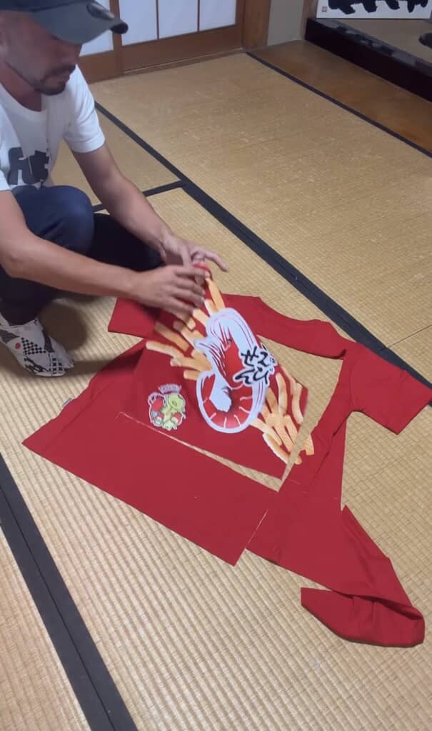 畳の上でカルビーかっぱえびせんTシャツをハサミで切り抜く男性