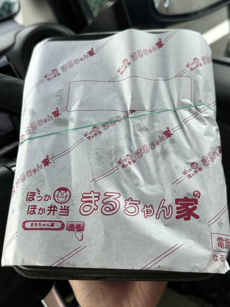 まるちゃん家（ほっかほか弁当）の包み紙に包まれた弁当を車内で持つ手