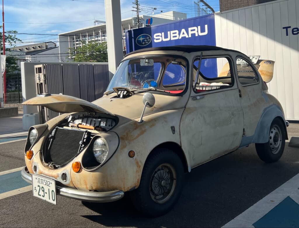 SUBARU販売店前に停まる旧型スバル360、ボンネットを開けたレストア途中の車体
