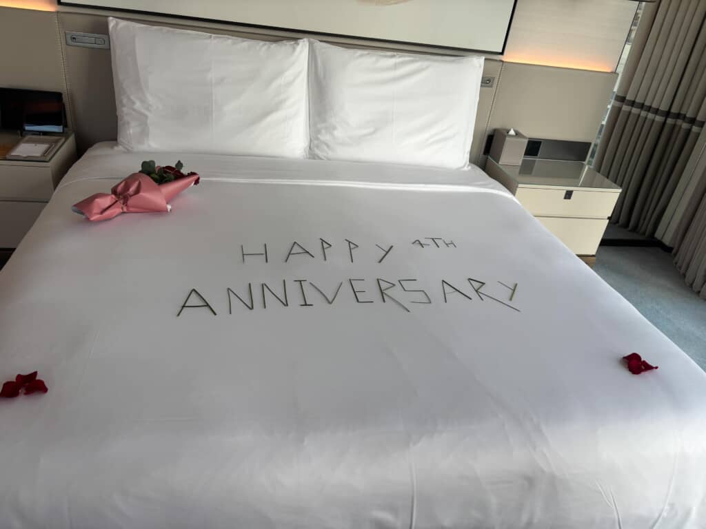 ホテルのベッドに「HAPPY 4TH ANNIVERSARY」の文字装飾と花束、バラの花びら